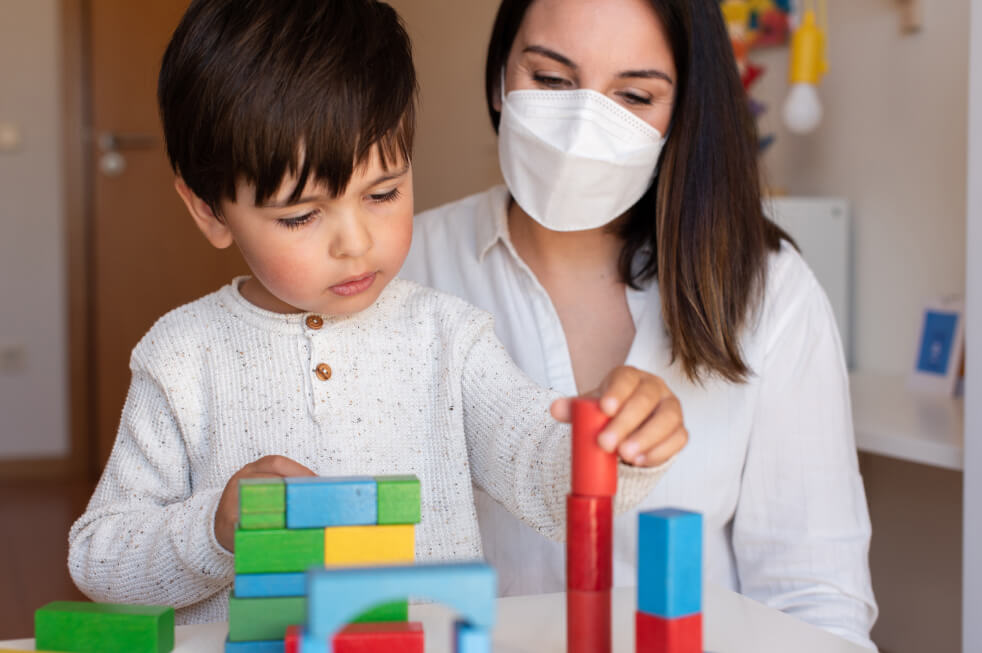 Masked teacher sitting with a child playing with colorful building blocks.