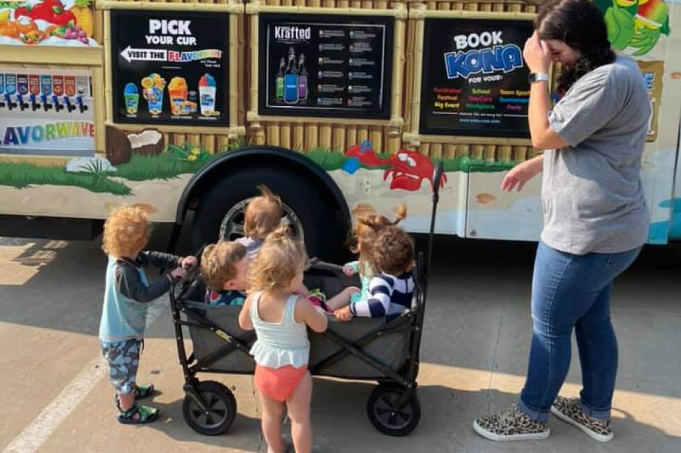 Teacher standing outside a Kona Ice truck with several toddlers standing at her feet.