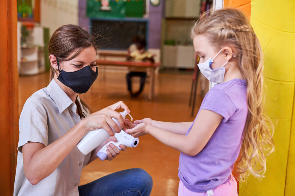Masked teacher squirting hand sanitizer onto a masked child's outstretched hands.