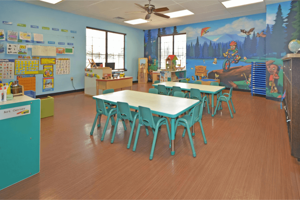 Childcare room with a mural on the back wall, blue tables and chairs, and toys along the walls.