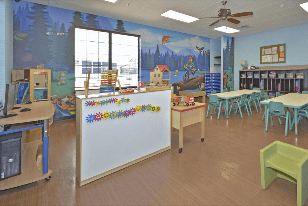 Childcare room with a mural on the back wall, two long tables and chairs, and a variety of toys around the room.