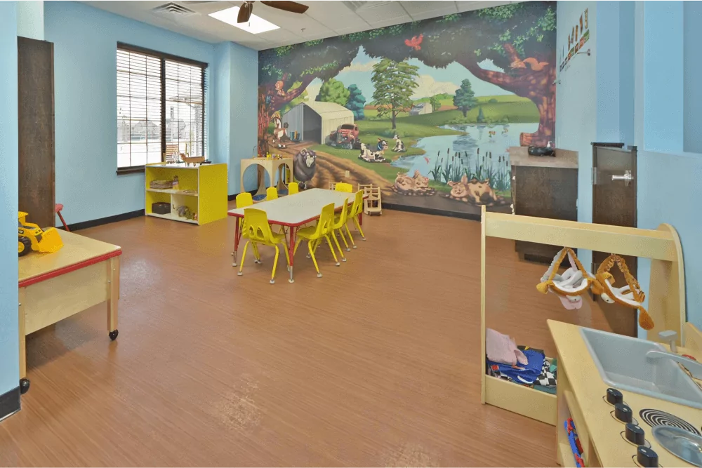 Toddler room with a forest and animal mural. The room has a table in the center with yellow chairs around it.