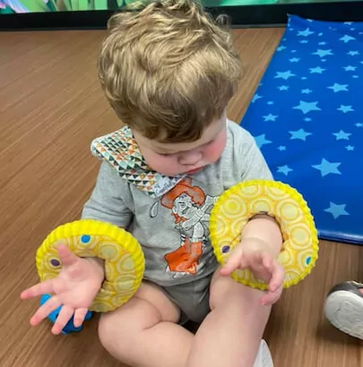 Blonde baby sitting on the floor with yellow donut-shaped toys around his wrists.