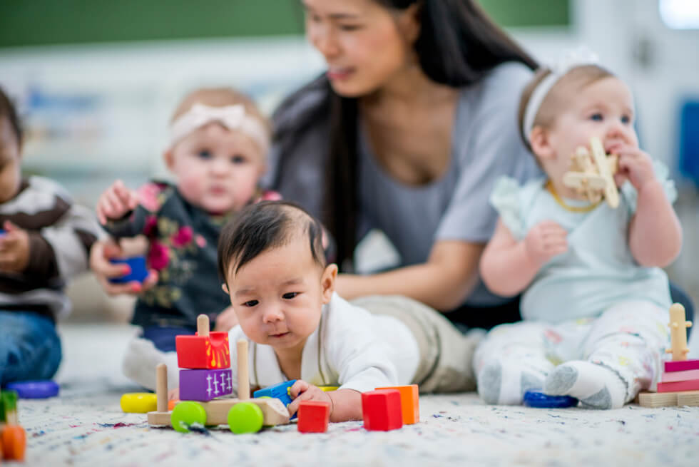 Infant lying on his stomach with blocks around him. In the background, a teacher is sitting on the floor next to other babies sitting up and playing with blocks.