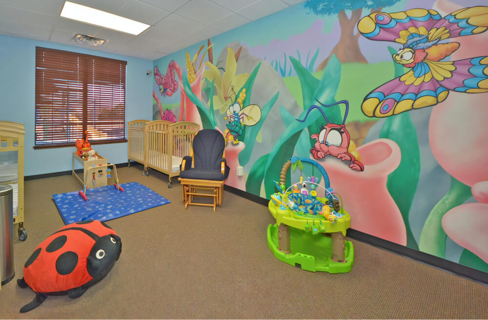 Classroom with a blue mat with a star pattern on the floor, cribs along the walls, a bright green bouncer, and a mural of insects and flowers on the wall.