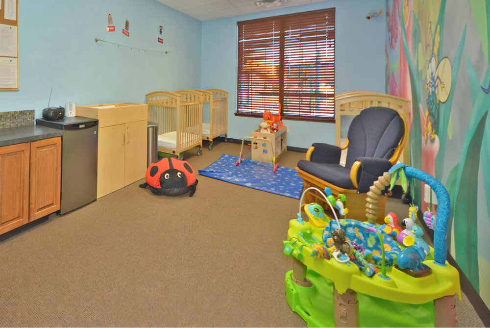 Classroom with a blue mat with a star pattern on the floor, cribs along the walls, a bright green bouncer, and a rocking chair.