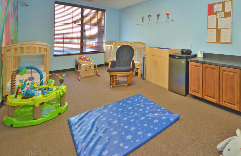 Classroom with a blue mat with a star pattern on the floor, cribs along the walls, a bright green bouncer, and a rocking chair.