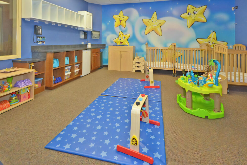 Classroom with two blue mats with star patterns on the floor, cribs by the wall, and a mural of clouds and smiling stars on the wall.