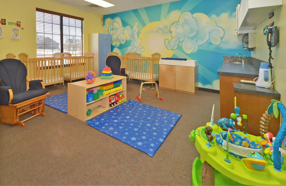 Classroom with two blue mats with star patterns on the floor, cribs by the wall, a rocking chair, a bright green bouncer, and a mural of clouds shaped like animals in front of the sun on the wall.