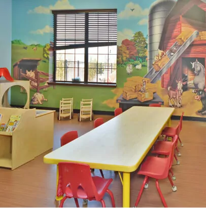Classroom with a long rectangular table with red plastic chairs around it. On the wall, there's a mural of a barn and barn animals.