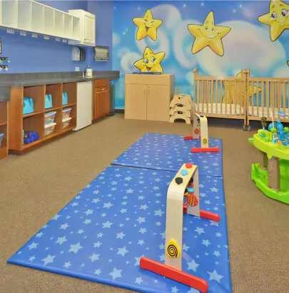 Classroom with two blue mats with star patterns on the floor, cribs by the wall, and a mural of clouds and smiling stars on the wall.