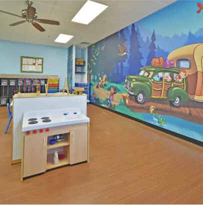 Classroom with a play kitchen set and a mural of a green car with a camper attached to the back parked next to a lake and the edge of a forest.