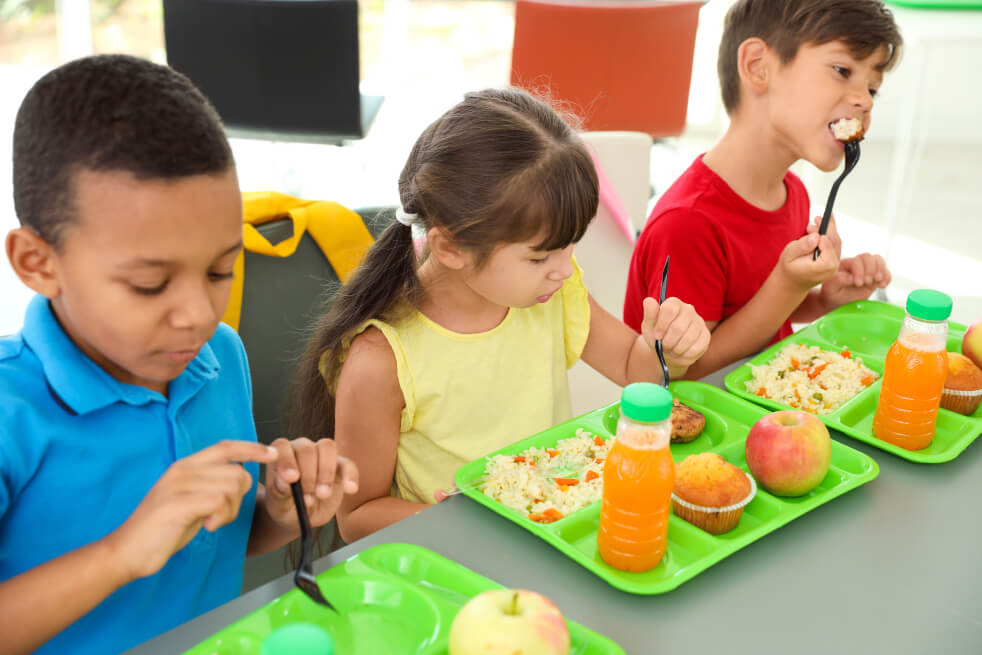 Young children sitting at a table with bright green trays of food in front of them.