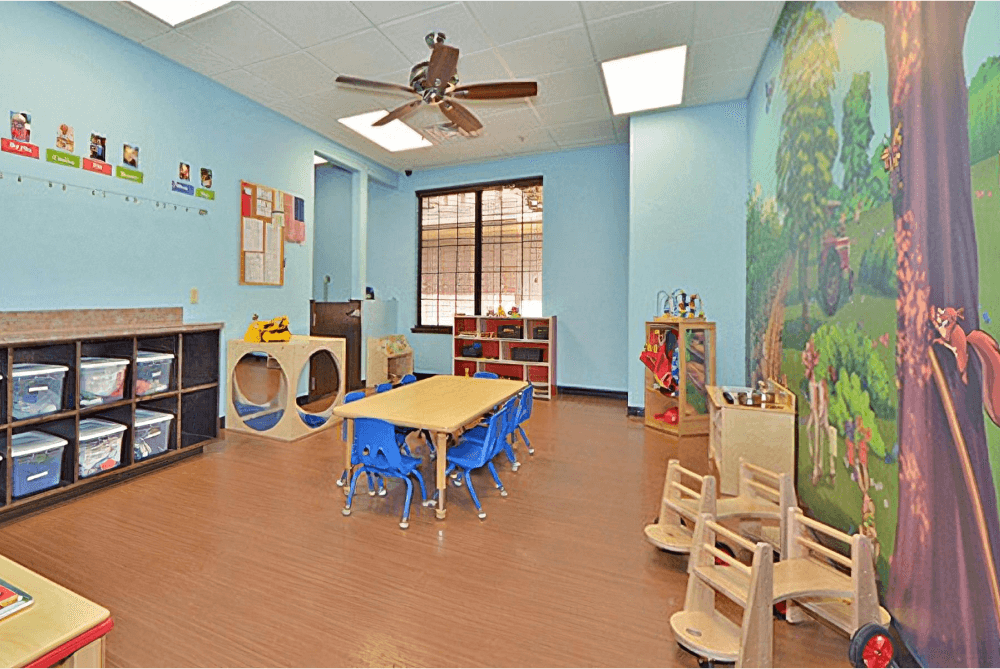 Childcare room with a forest mural, a long table with blue chairs, containers full of toys, and other toys around the edges of the room.