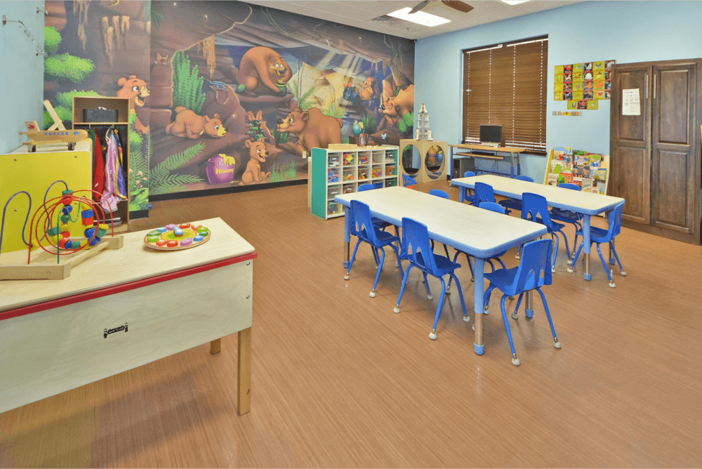 Childcare room with a mural of bears, two long tables with blue chairs, and a table with toys on it.