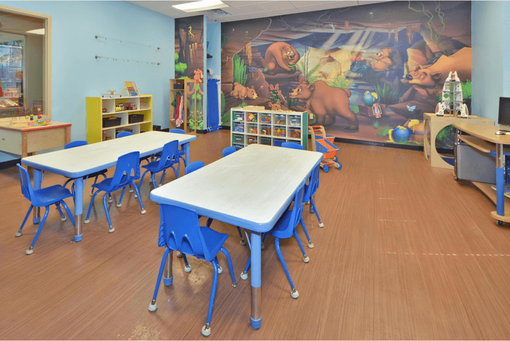 Childcare room with a bear mural, two long tables with blue chairs, and toy containers along the walls.