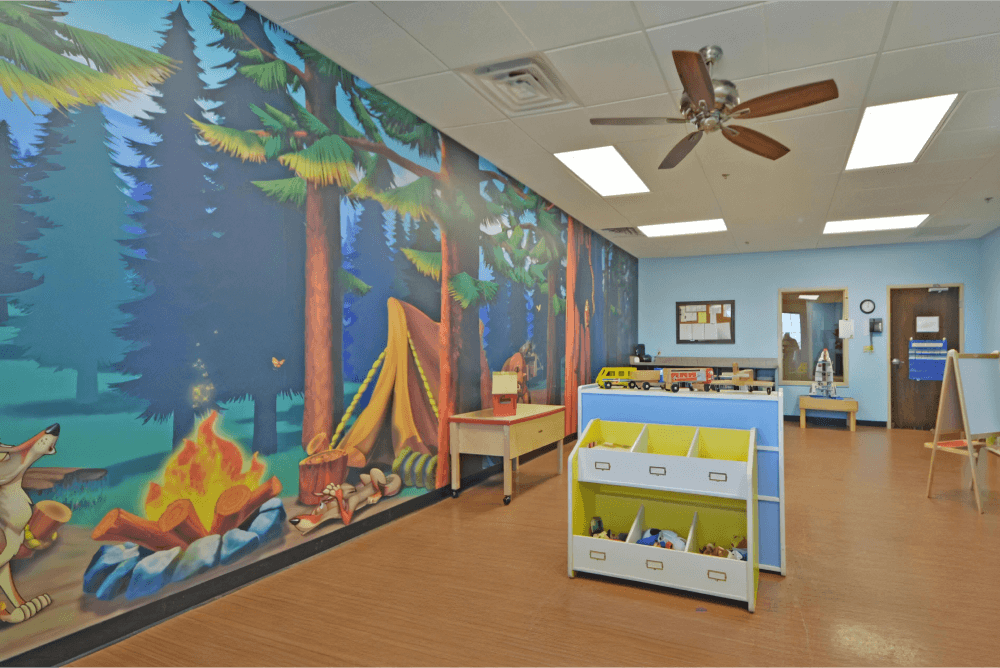 Childcare room with a forest mural, multiple containers with toys, and an easel in the background.