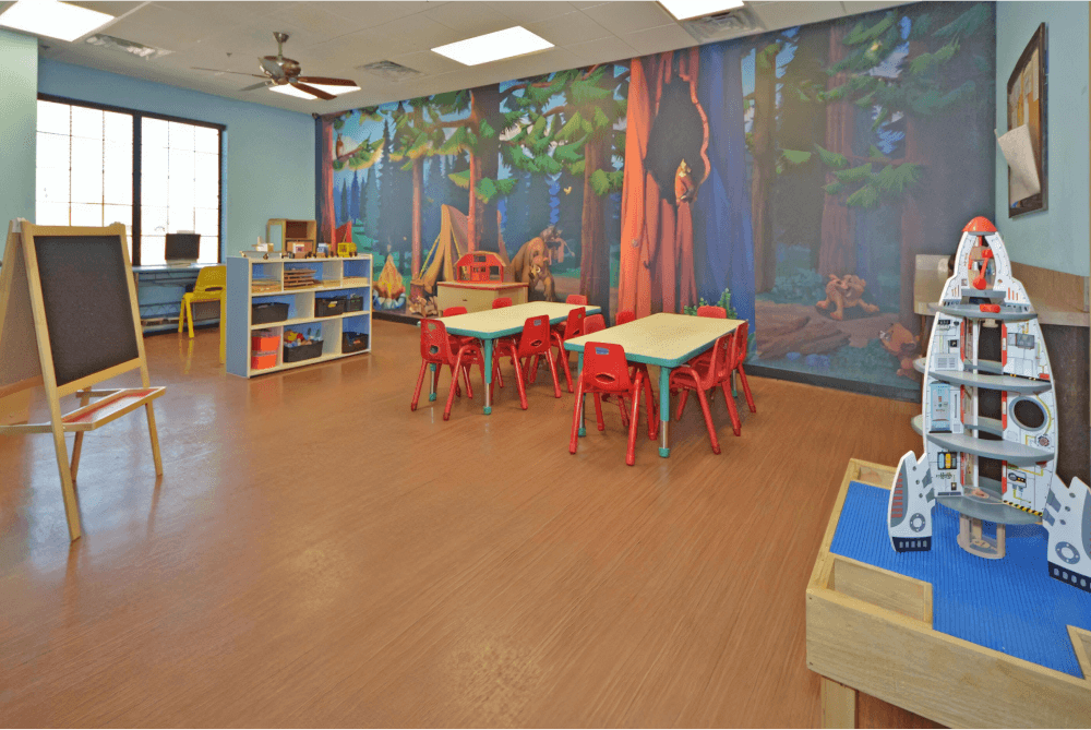 Childcare mural with a forest mural, two long tables with red chairs, and an easel on the left side of the room.