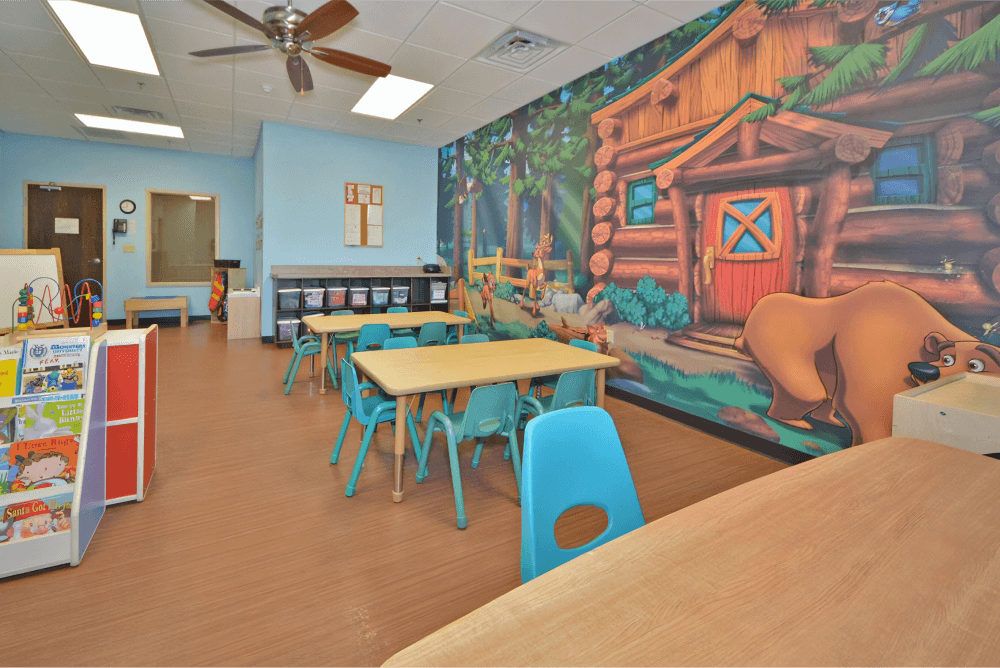 Childcare room with a cabin mural on the wall, multiple tables with blue chairs, and a bookshelf in the background.