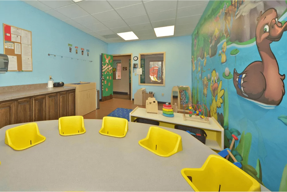 Toddler room with circular table with yellow highchairs built in. There is a other toys and a pond mural in the background.