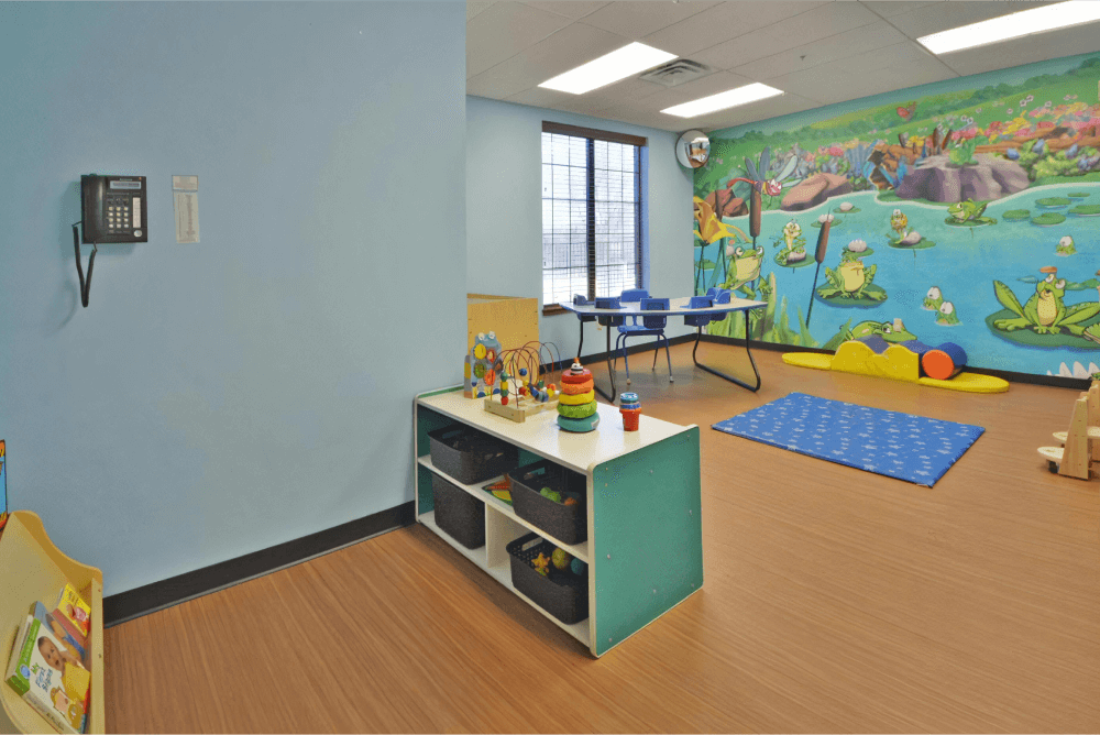 Toddler room with a pond mural on the wall. There is storage containers with toys inside and a blue mat in the center of the floor.