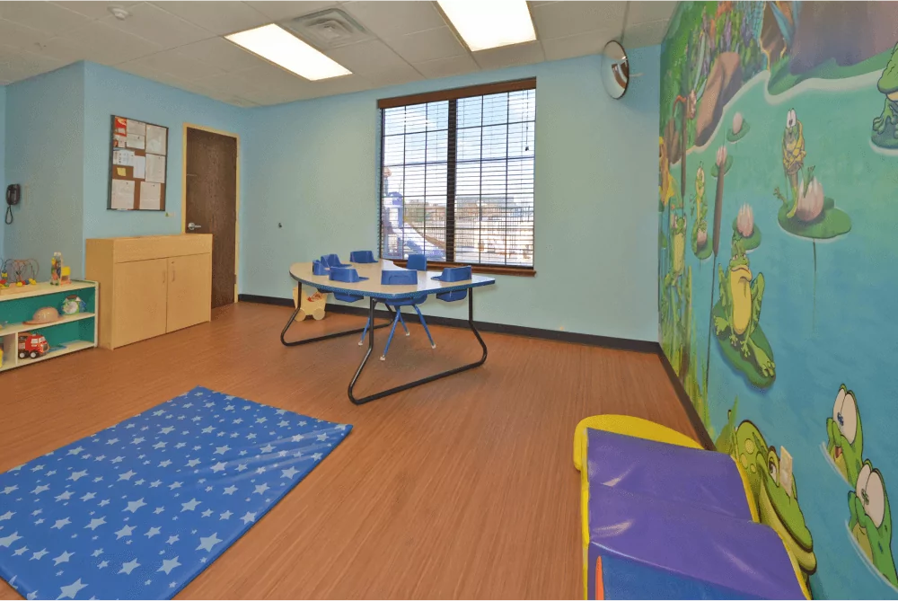 Toddler room with a pond mural, a blue mat on the floor, and a table with built in high chairs.