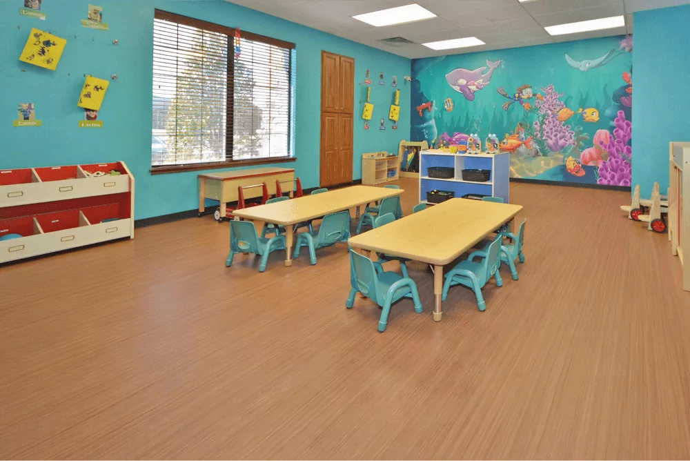 Toddler room with blue walls and an ocean mural. There are two long tables in the center with blue chairs around them.