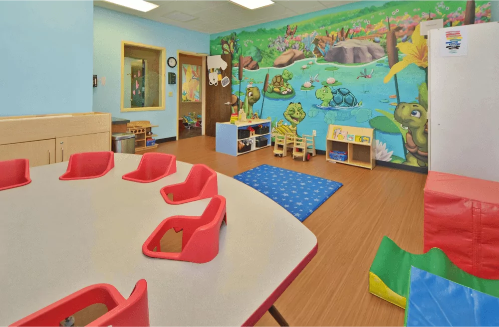 Toddler room with pond mural on the back wall. There is a table with built in high chairs and a blue mat on the floor.