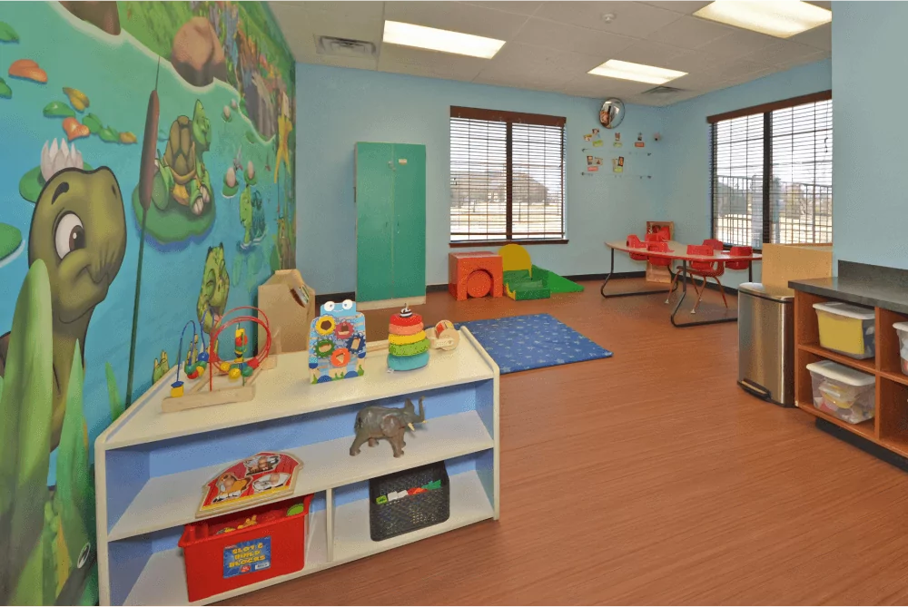 Toddler room with a pond mural on the wall. There are storage containers with toys around the room and there is a table in the back corner.