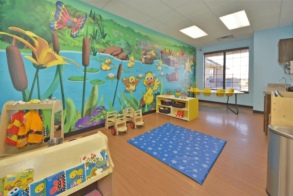 Toddler rooms with a pond mural on the back wall. There are toys throughout the room, a blue mat in the center of the room, and a table with yellow highchairs next to a window.