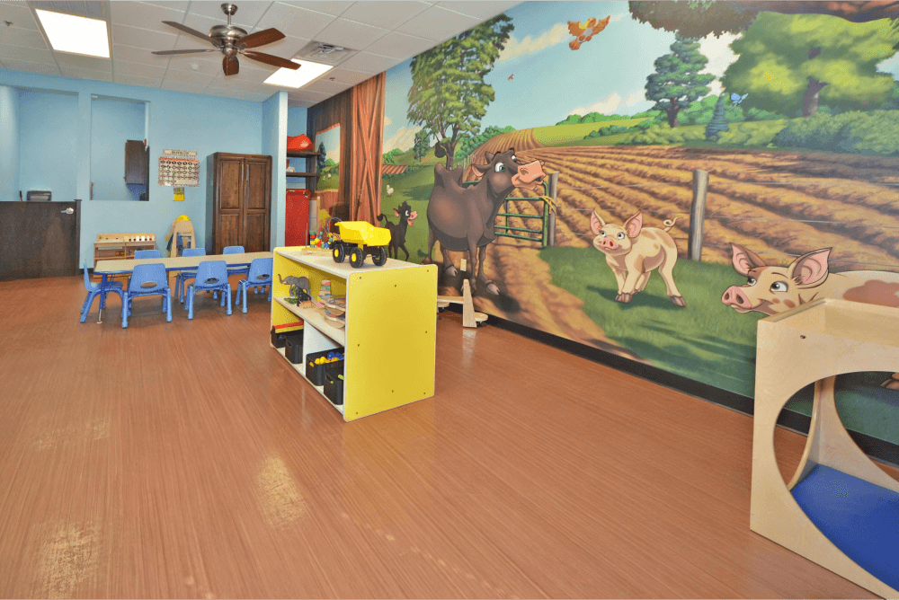 Childcare room with farm mural, a yellow toy storage container, and a table with blue chairs.