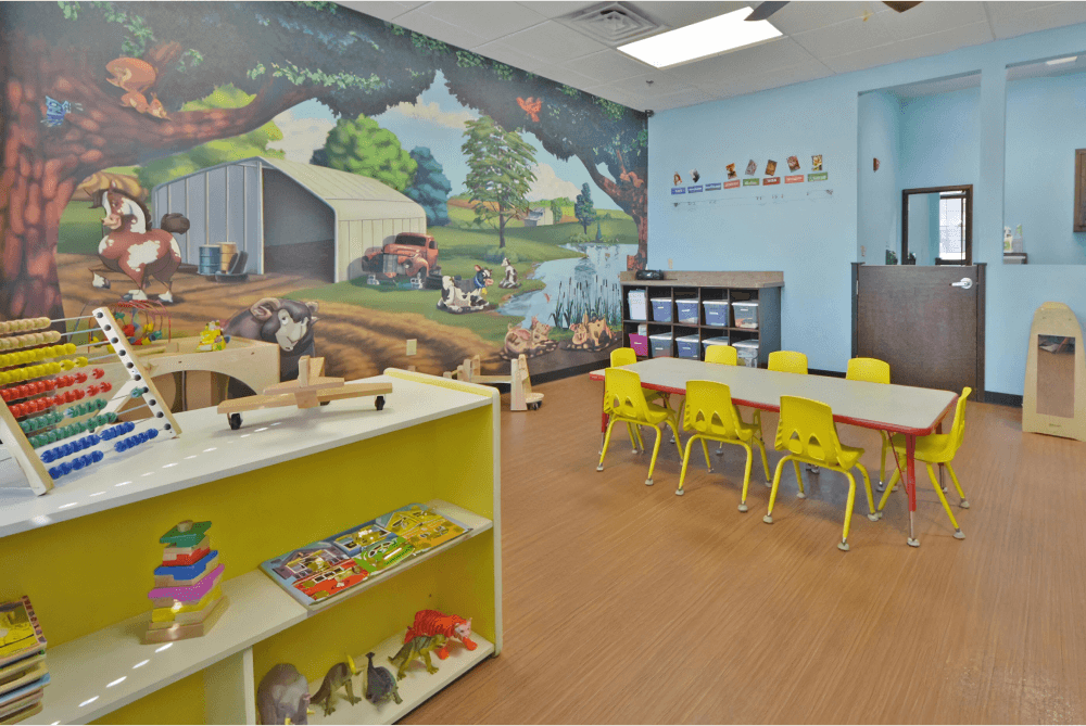 Childcare room with a farm animal mural, blue walls, a yellow toy storage shelf, and a table with yellow chairs.