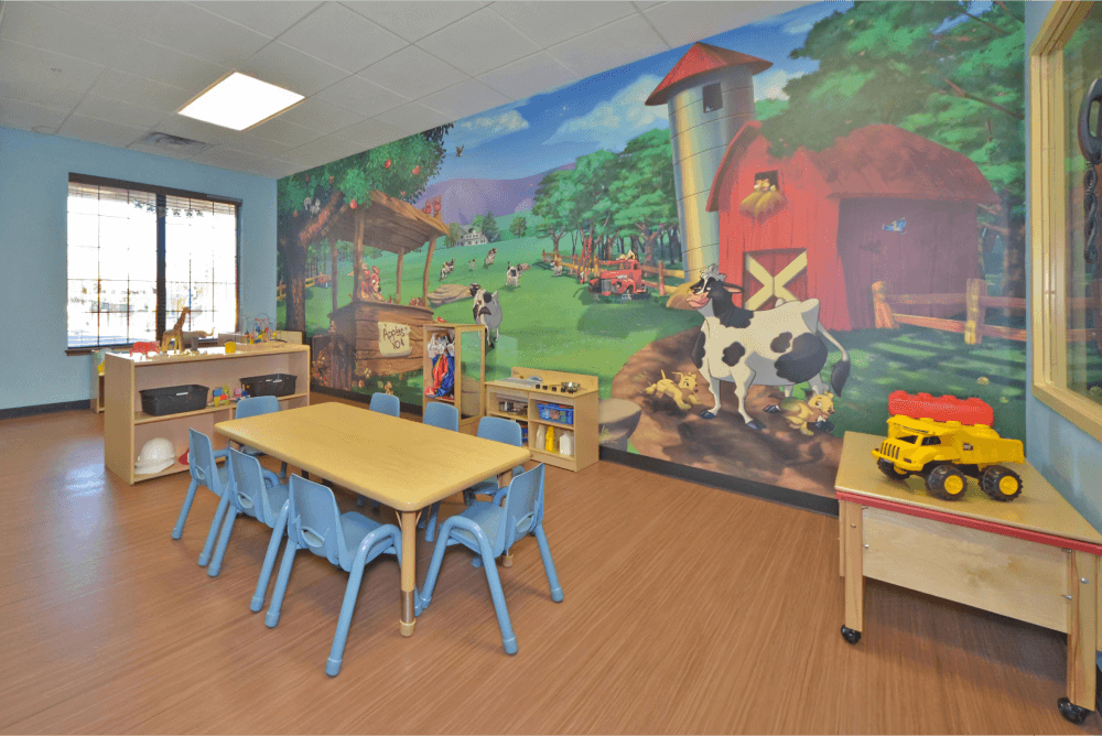 Childcare room with a farm animal mural, blue walls, toy storage shelfs, and a table in the center with blue chairs.