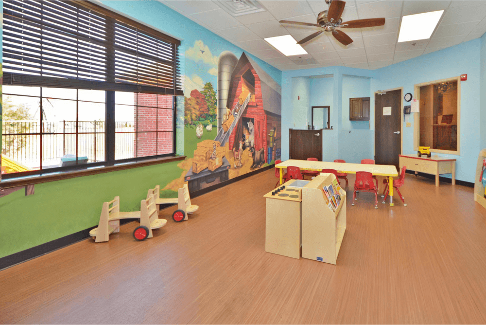 Childcare room with a farm mural, blue wall, a table with red chairs, and toys in the center of the room.