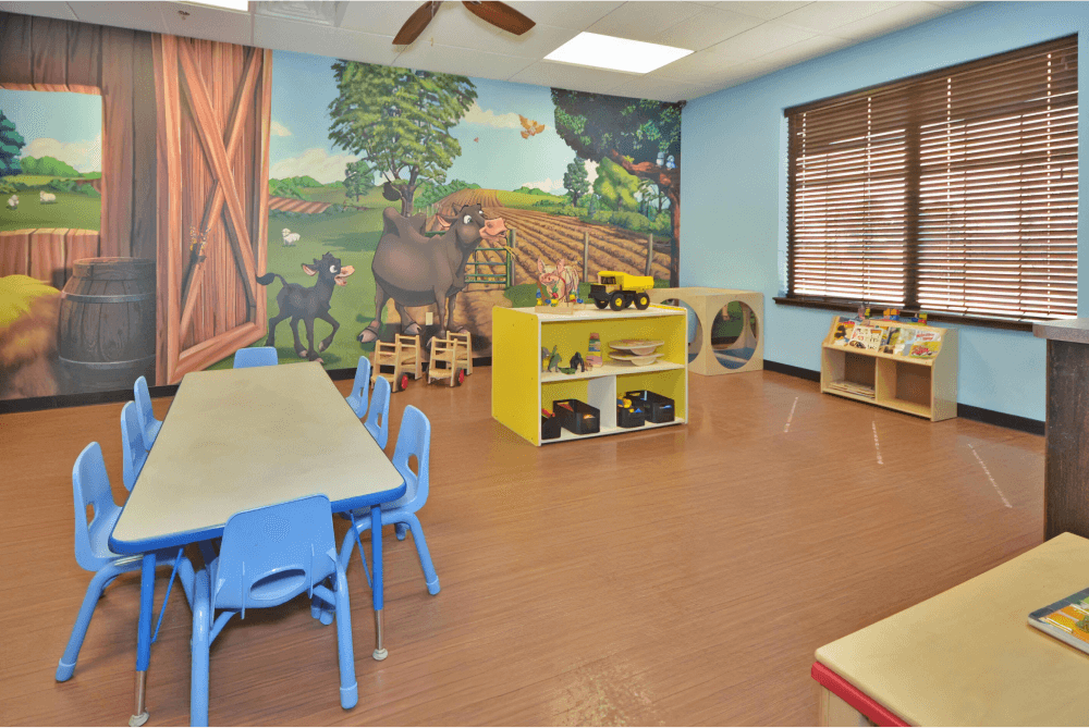 Childcare room with a farm mural, toy storage containers, and a table with blue chairs.