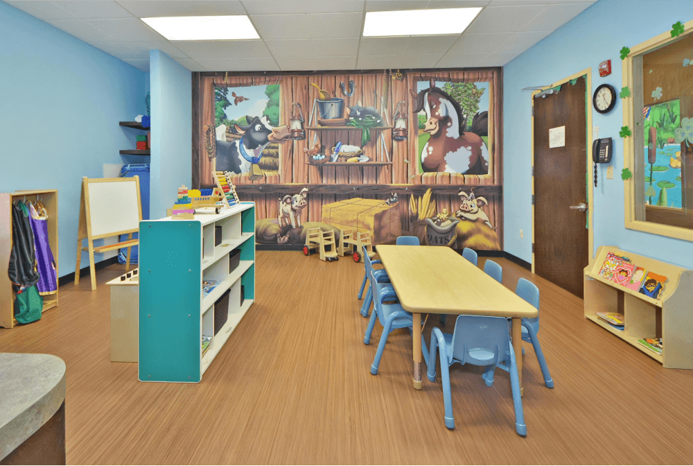 Childcare room with a farm animal mural, a table with blue chairs in the center, and toys around the edge of the room.