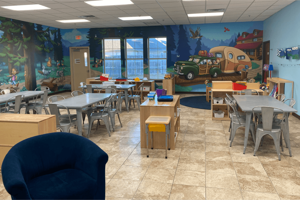 classroom with a campsite mural, multiple tables and chairs, and cubbies with supplies in them.