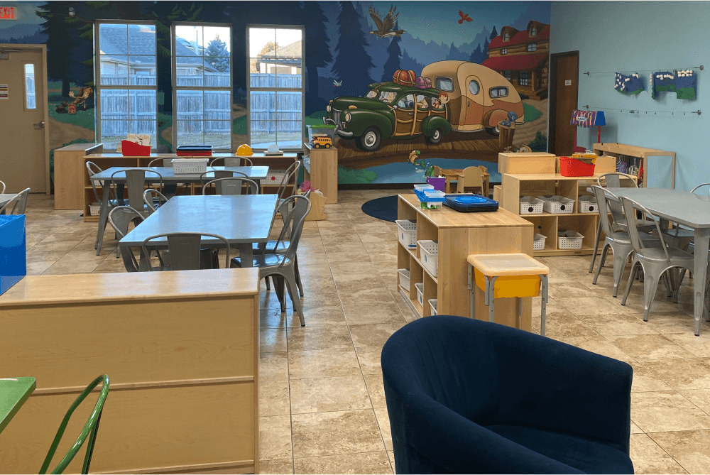 Classroom with a campsite mural, blue walls, long tables and chairs, a navy blue chair, and cubbies with various supplies in them.