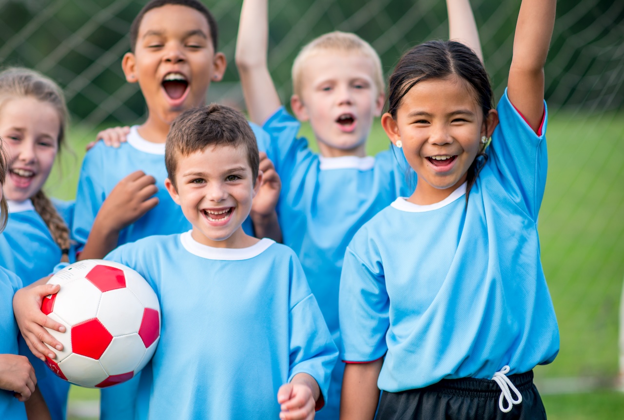 A small group of elementary aged children cheer after a soccer match as they celebrate their win. They are each wearing a blue jersey and are smiling.