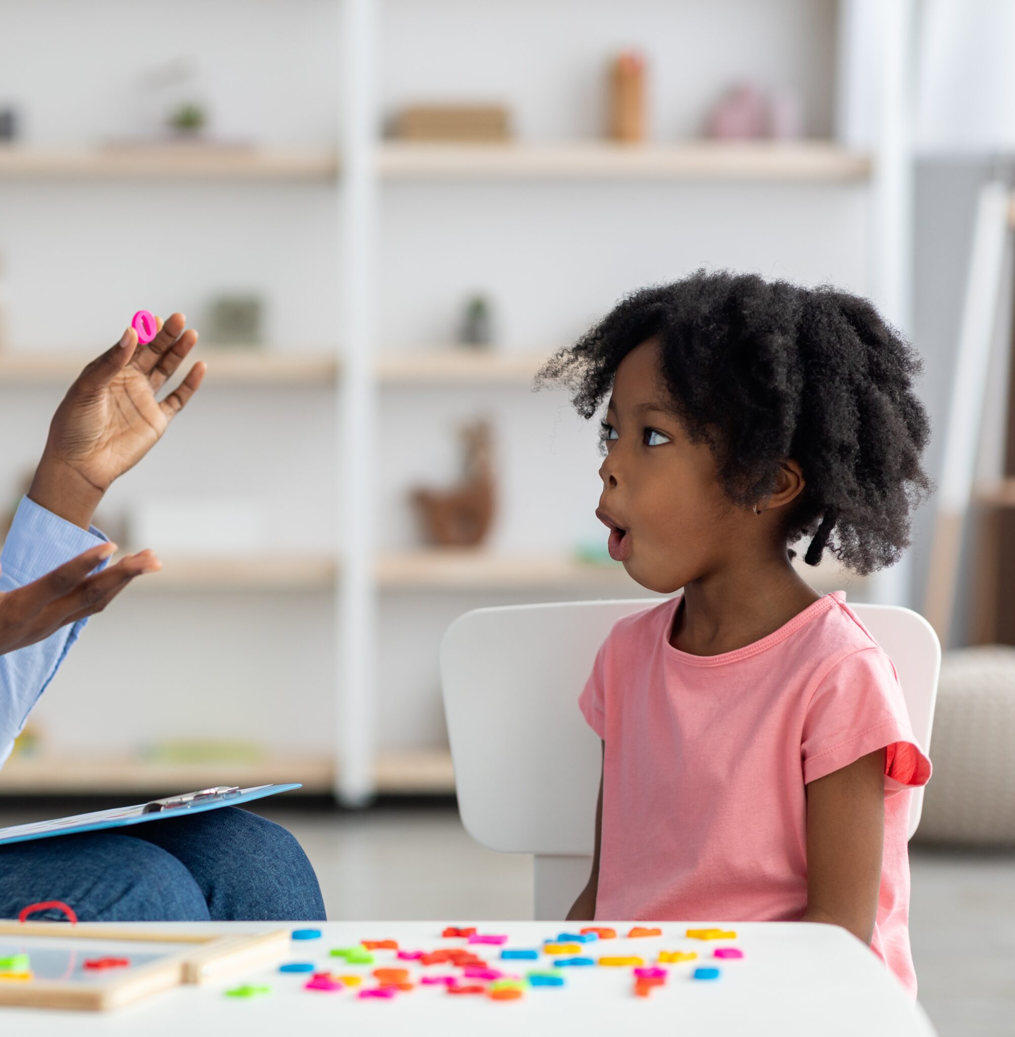 Attractive young african american woman speech therapist working with little black girl, sitting at table with colorful letters, learning pronunciation, clinic interior. Speech disorders in children